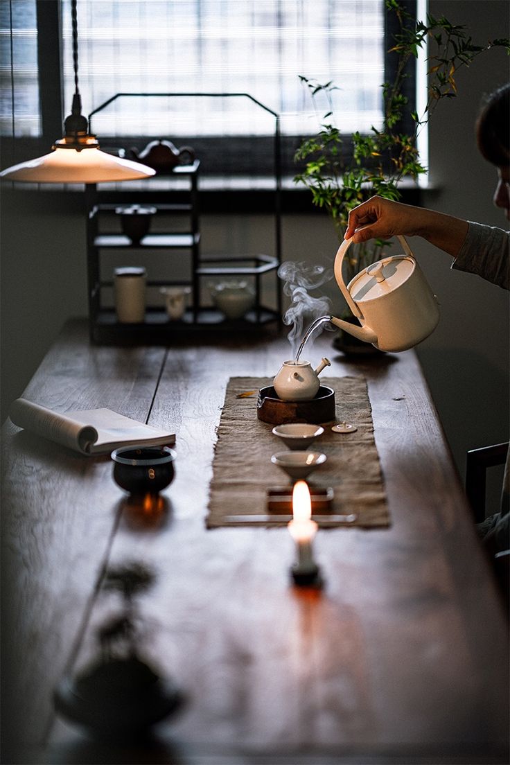 a woman pours tea from a pitcher into a cup on a table with candles