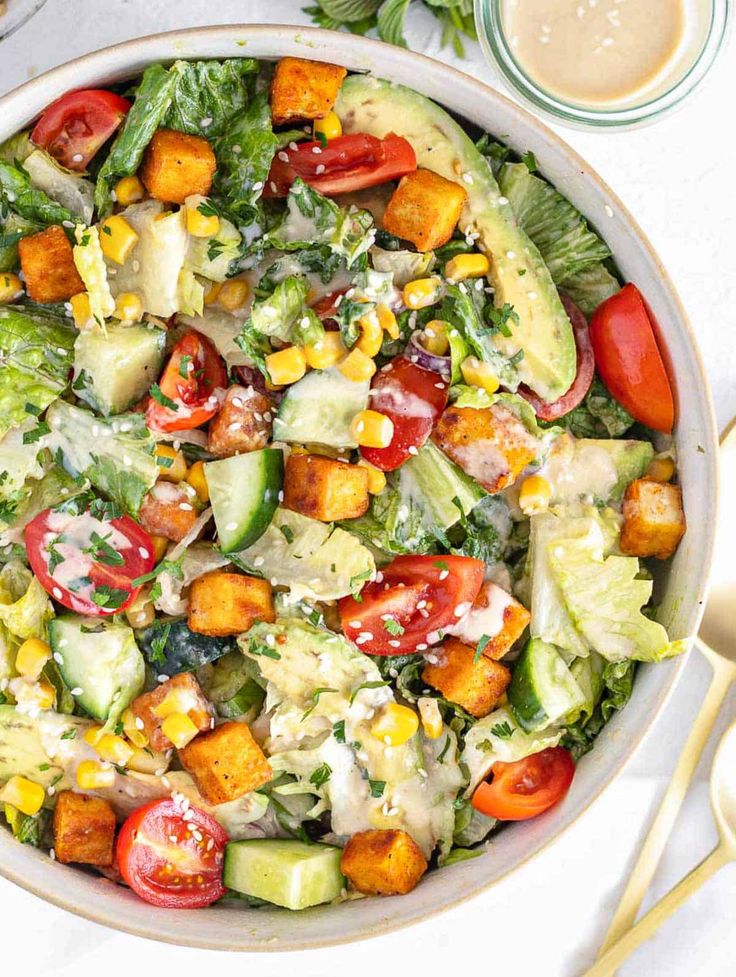 a salad in a bowl with dressing next to it on a white tablecloth and utensils