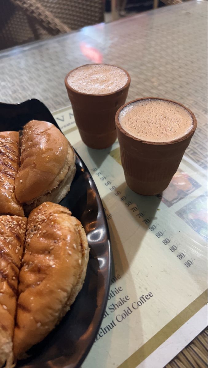 two cups of coffee sit on a table next to pastries and sandwiches, which are sitting on a plate