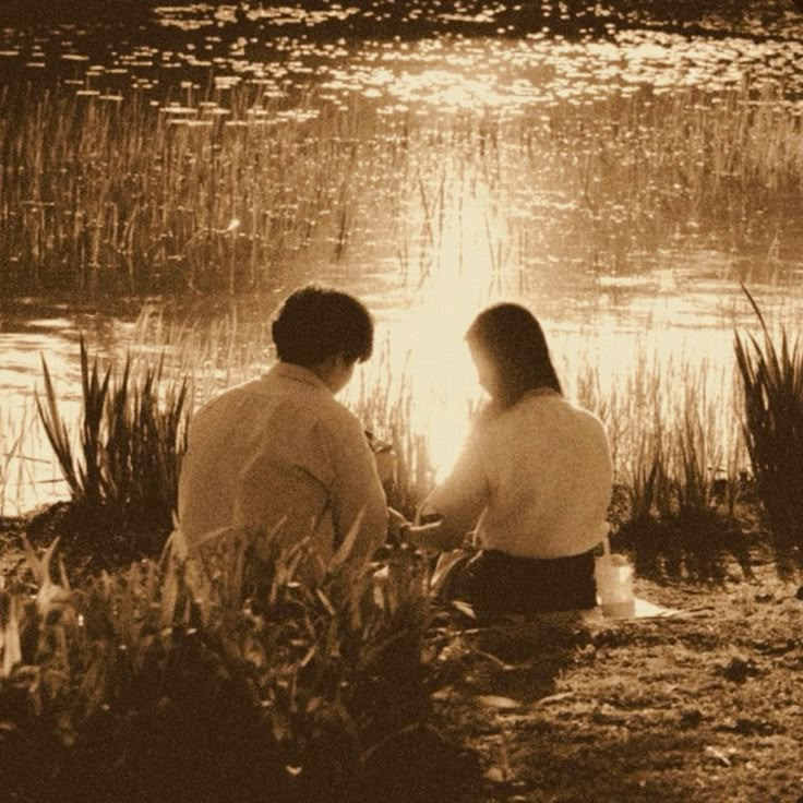 two people sitting on the shore of a lake in front of tall grass and reeds