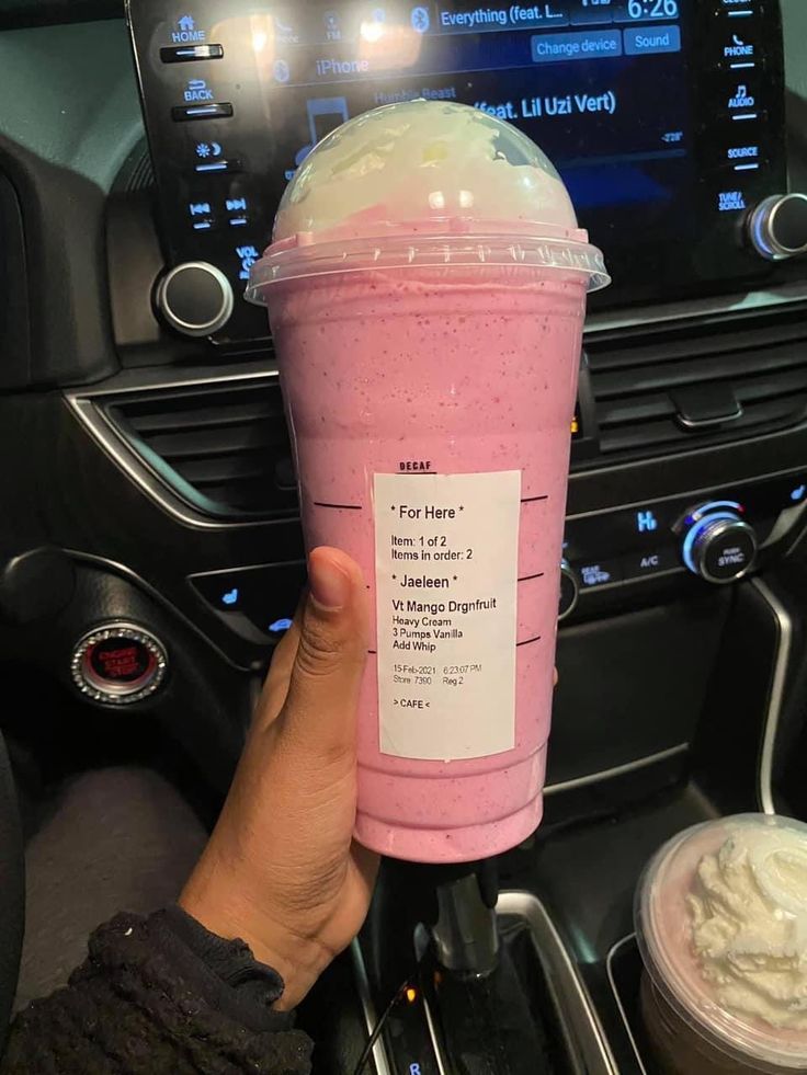 a person holding up a pink drink in their hand while sitting on the car dashboard