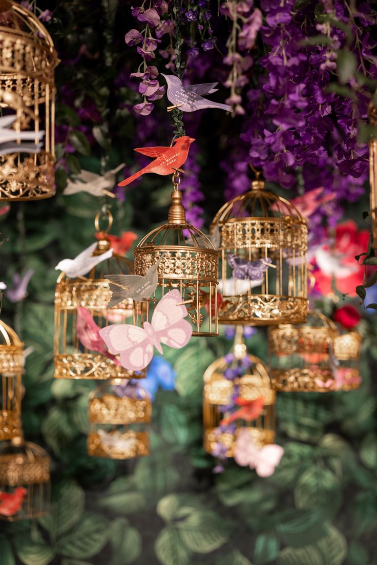 several birdcages hanging from the ceiling in front of purple and green flowers with birds flying around them