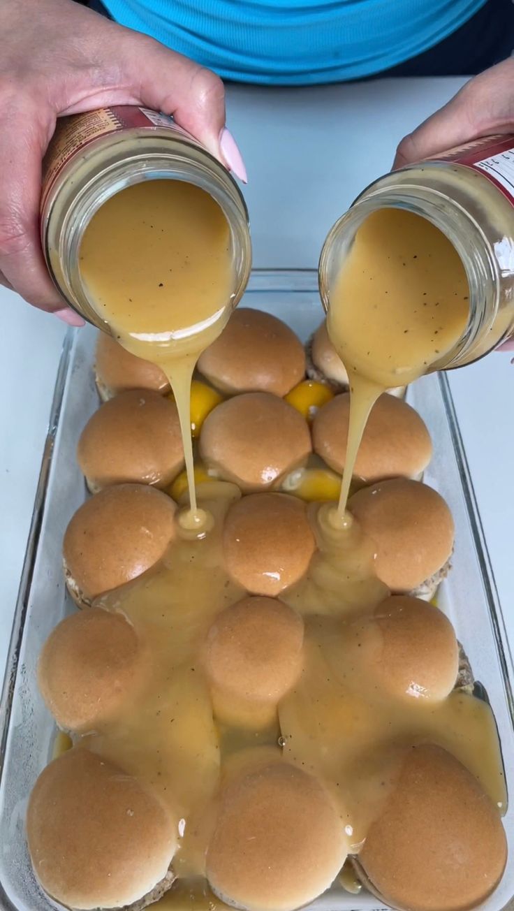 two hands pouring sauce over mini hamburgers in a baking dish with buns on the side