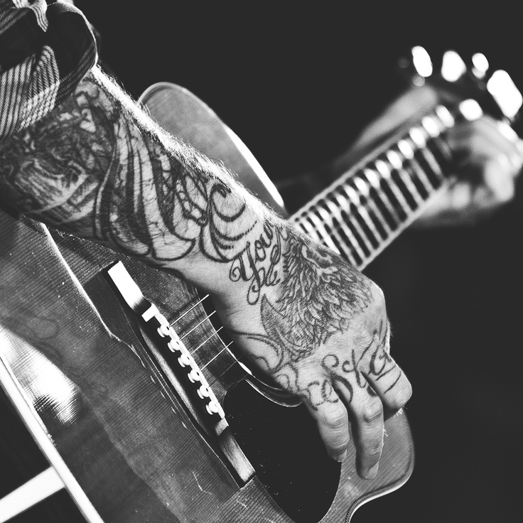 black and white photograph of a man with tattoos on his arm playing an acoustic guitar