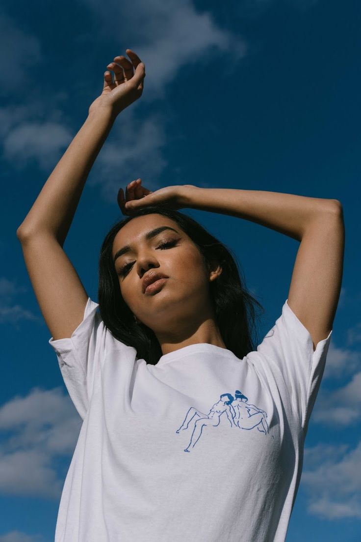 a woman in white shirt holding her hands up to her head with blue sky and clouds behind her