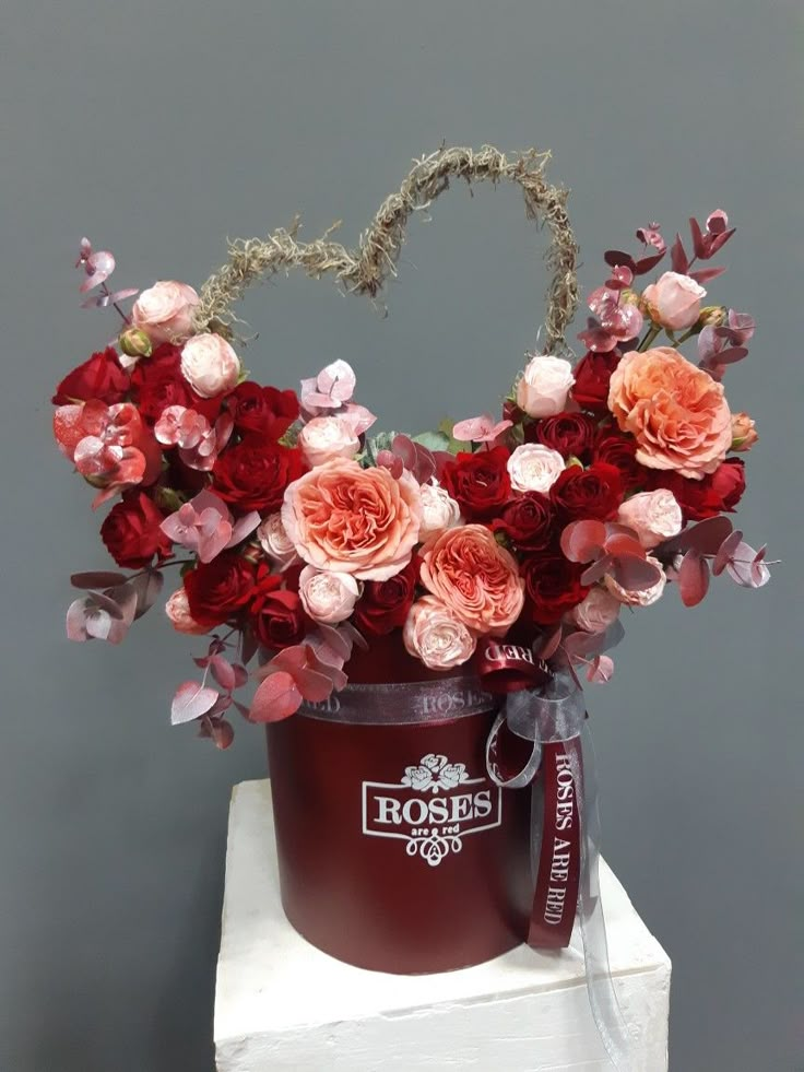 a red bucket with pink flowers and greenery in it sitting on a white pedestal