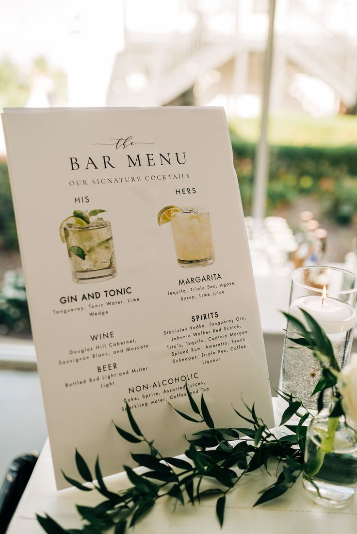 a menu with drinks on it sitting next to a glass filled with water and greenery