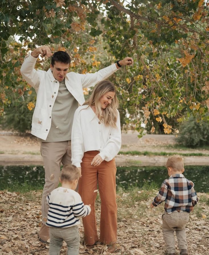 a man, woman and two children standing in front of a tree with leaves on the ground