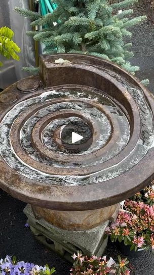 an outdoor fountain with flowers around it and a tree in the back ground behind it