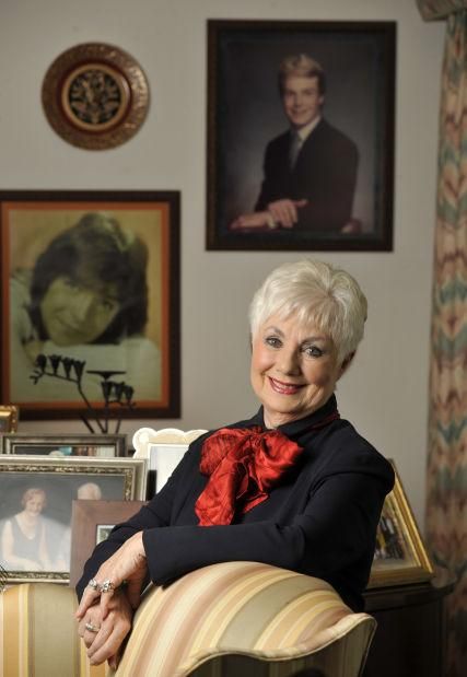 an older woman sitting in a chair with pictures on the wall behind her and smiling at the camera