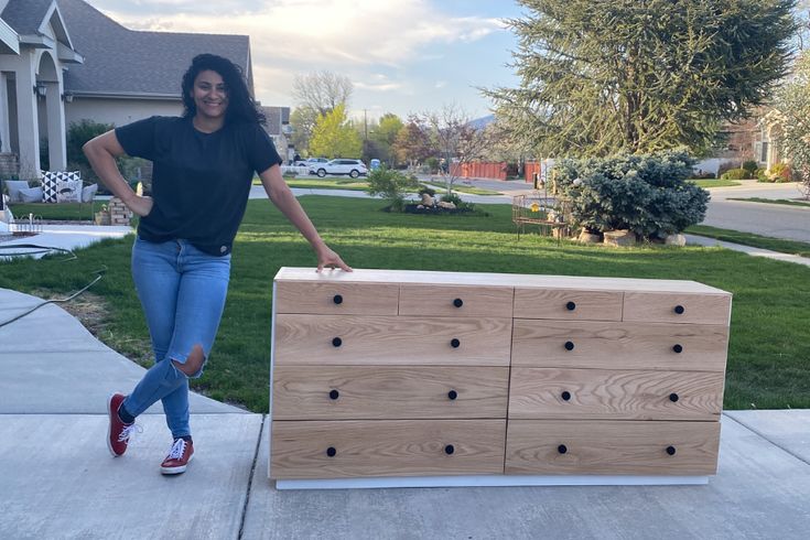 a woman standing next to a wooden dresser on the sidewalk in front of a house