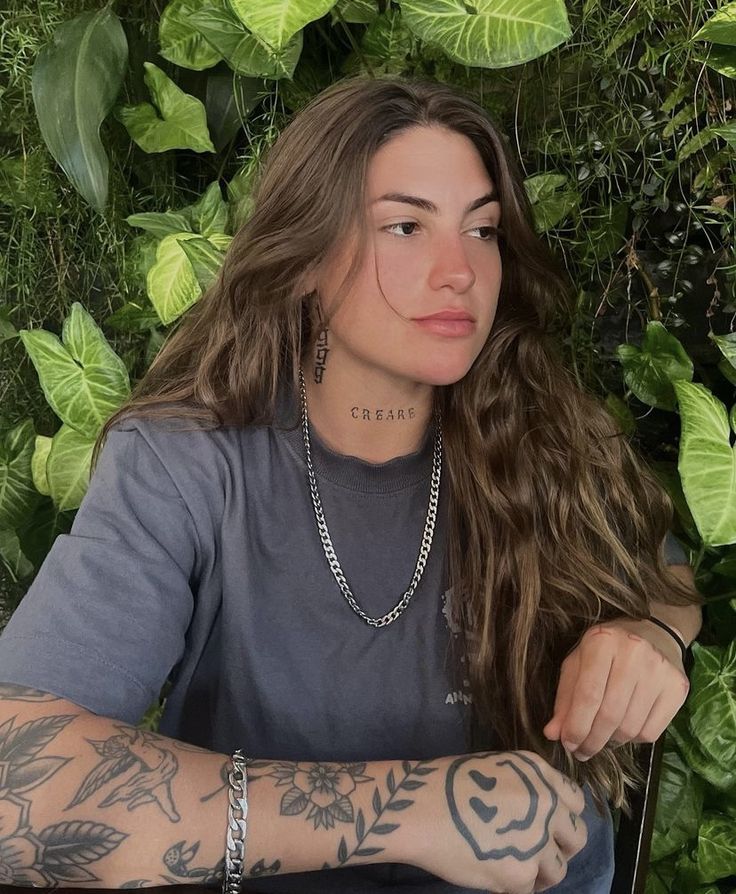 a woman with long hair and tattoos sitting in front of greenery, looking at the camera