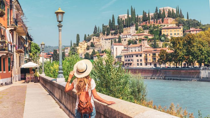 a woman wearing a hat is looking at the water and buildings in the background with a lamp post