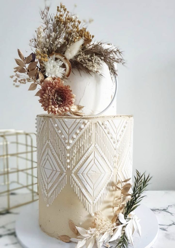 a white and gold wedding cake decorated with dried flowers, leaves and feathers on a marble table