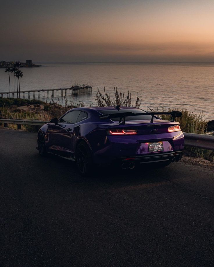 a purple sports car is parked on the side of the road by the ocean at sunset