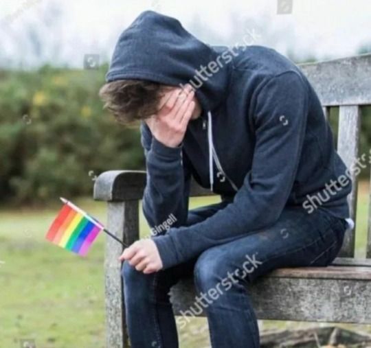 a man sitting on a bench with his head down holding a rainbow flag in front of him