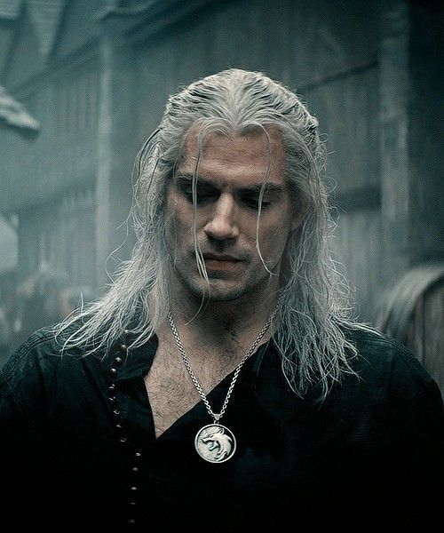 a man with long white hair wearing a black shirt and silver necklace standing in front of a building