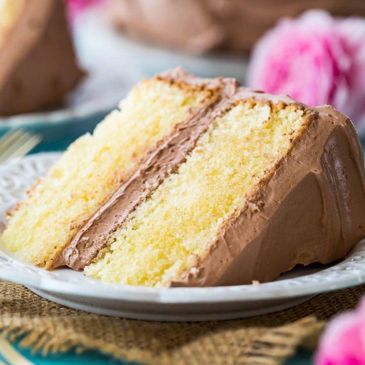 a slice of cake on a plate with chocolate frosting and flowers in the background