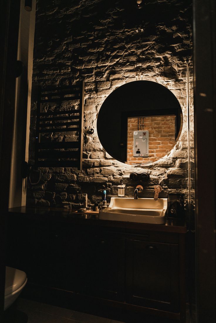 a bathroom sink under a round mirror in a dark room with brick walls and floor