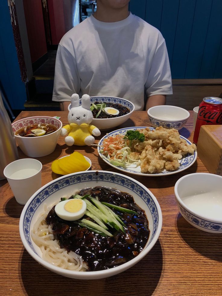 a person sitting at a table with many plates of food on it and bowls of soup in front of them