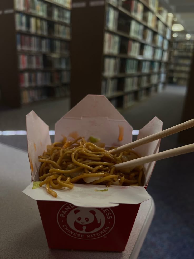 two chopsticks sticking out of a box filled with noodles in front of bookshelves