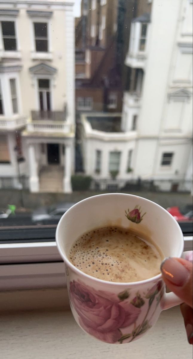 a person holding a cup of coffee in front of a window with buildings behind it