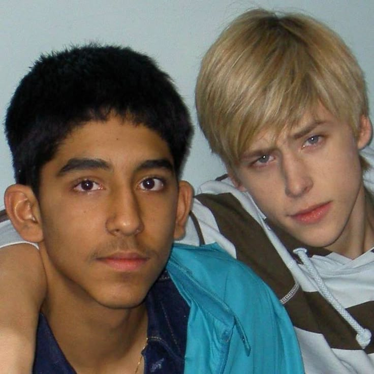 two young men sitting next to each other in front of a white wall and one is looking at the camera