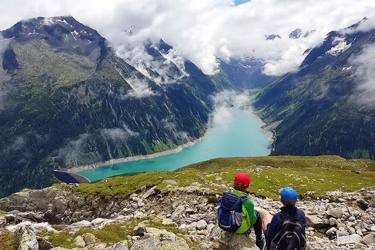 two people with backpacks looking out over a valley and lake in the mountains on a cloudy day