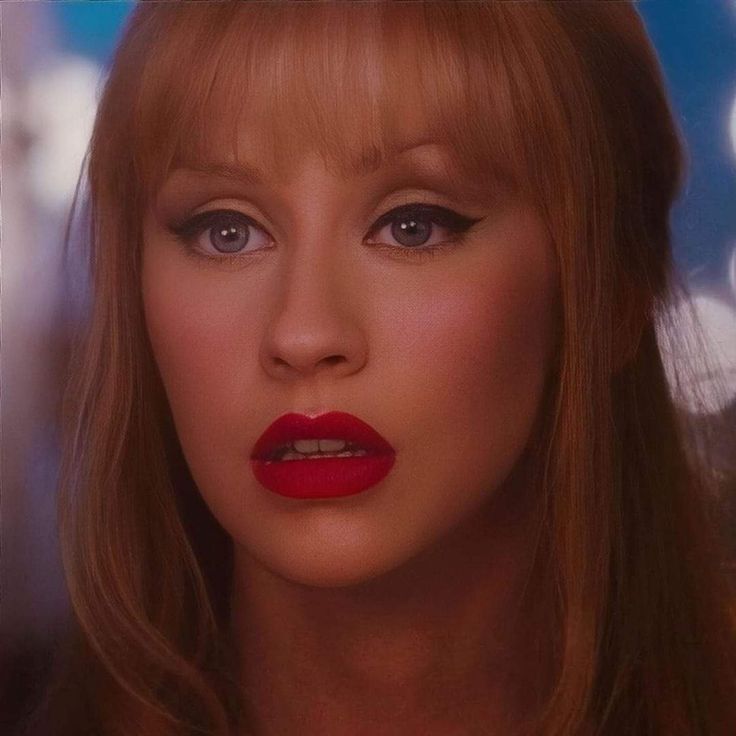 a woman with long blonde hair and blue eyes looks into the distance while wearing red lipstick