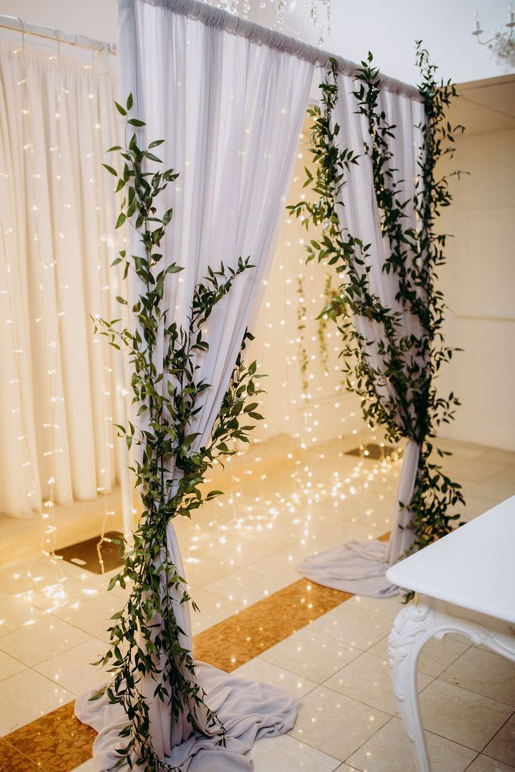 an indoor area with white curtains, lights and greenery on the wall behind it