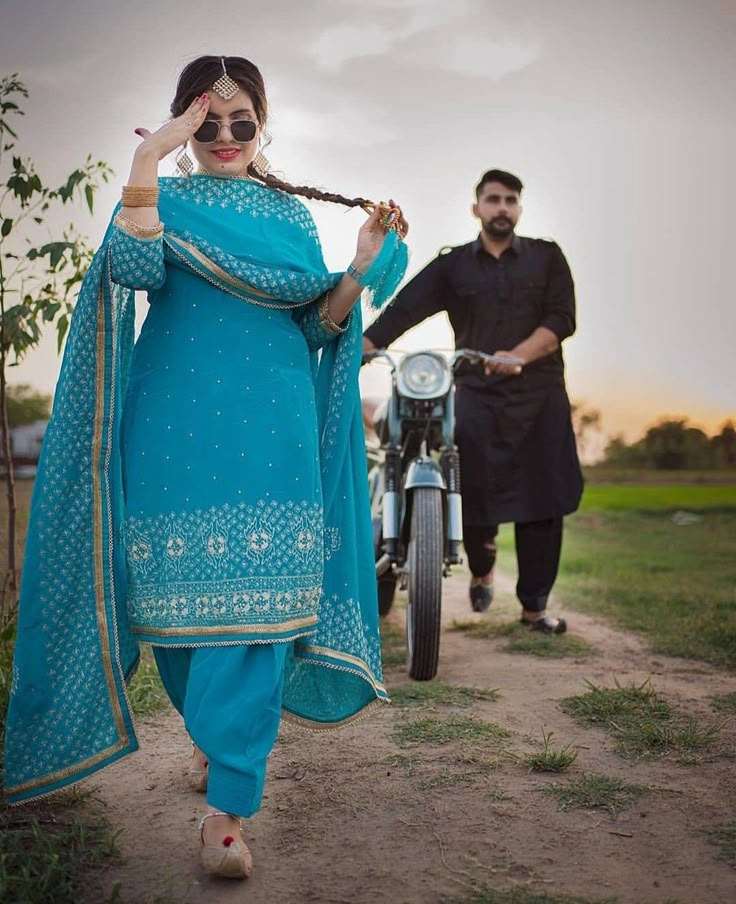 a man and woman walking down a dirt road next to a motorcycle in the background