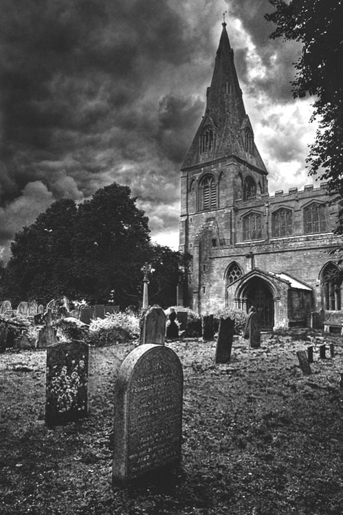 black and white photograph of an old church with tombstones in the foreground on a cloudy day