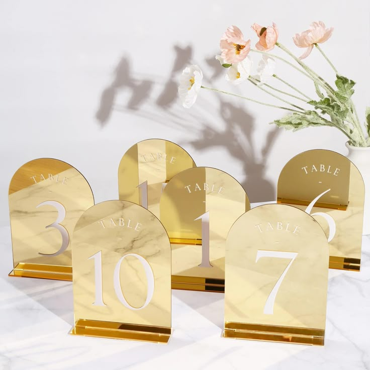 three gold table numbers sitting next to a vase with flowers in it on a marble surface