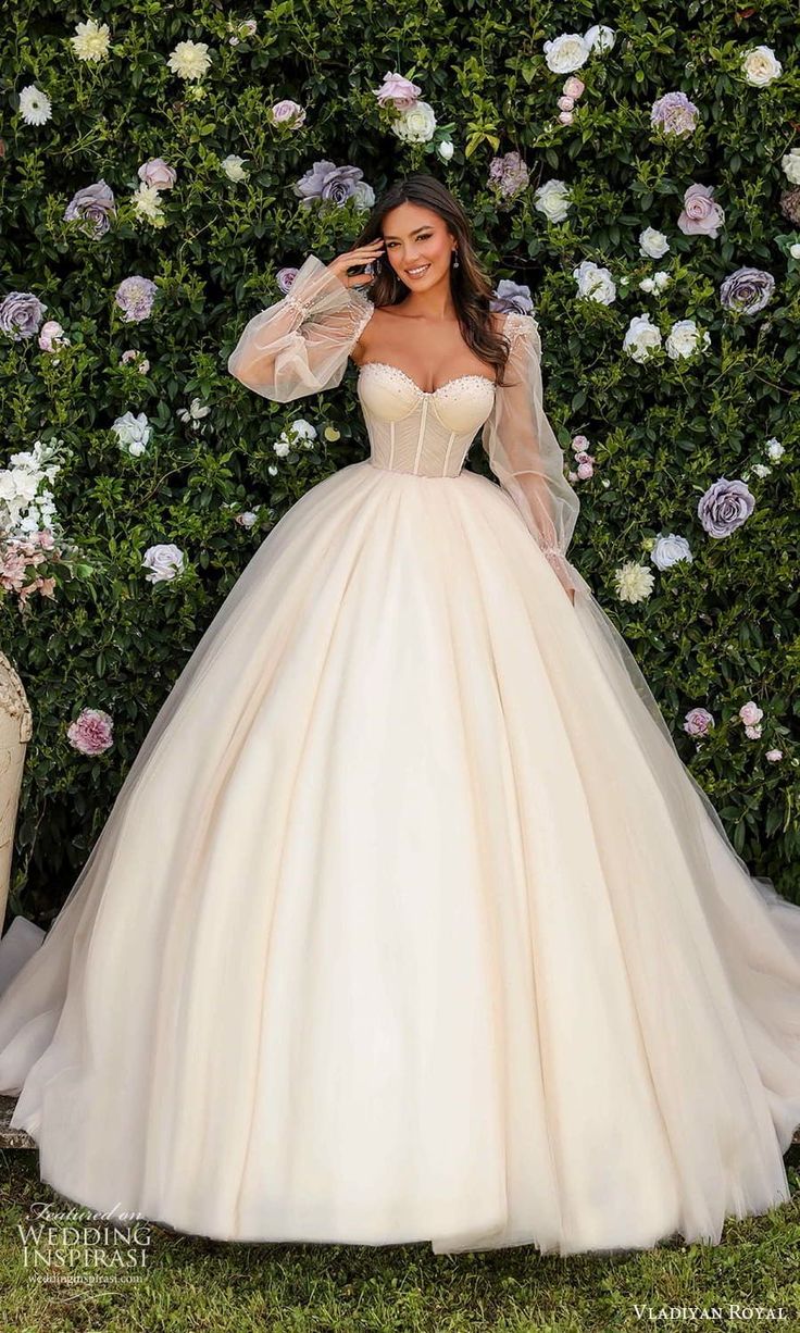 a woman standing in front of a flower covered wall wearing a wedding dress with long sleeves