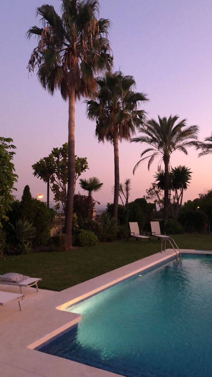 an empty swimming pool with lounge chairs and palm trees in the background at dusk or dawn