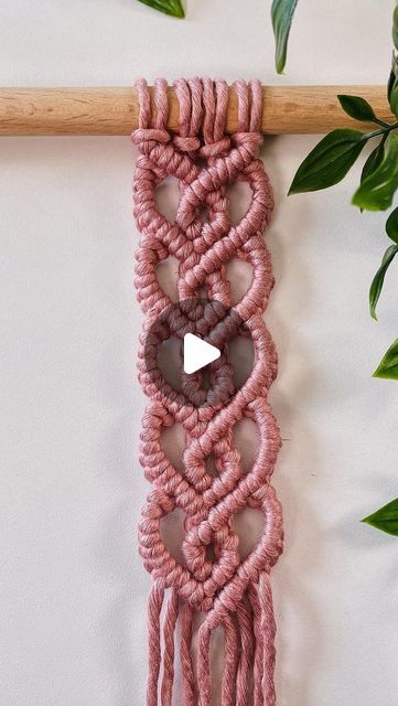 a pink macrame hanging on a wall next to a plant with green leaves