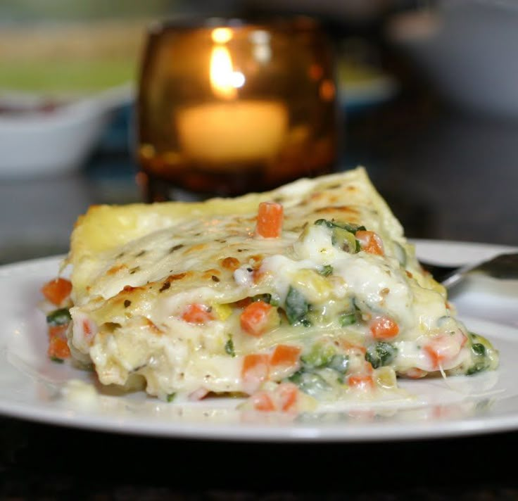 a slice of vegetable lasagna on a white plate with a candle in the background