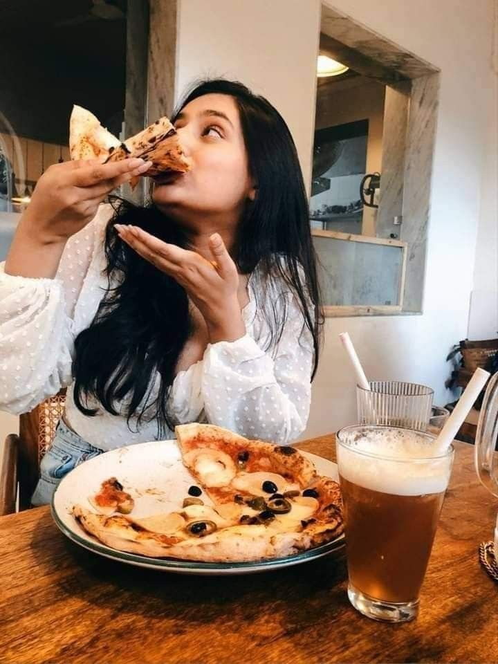 a woman sitting at a table eating pizza and drinking beer in front of her face