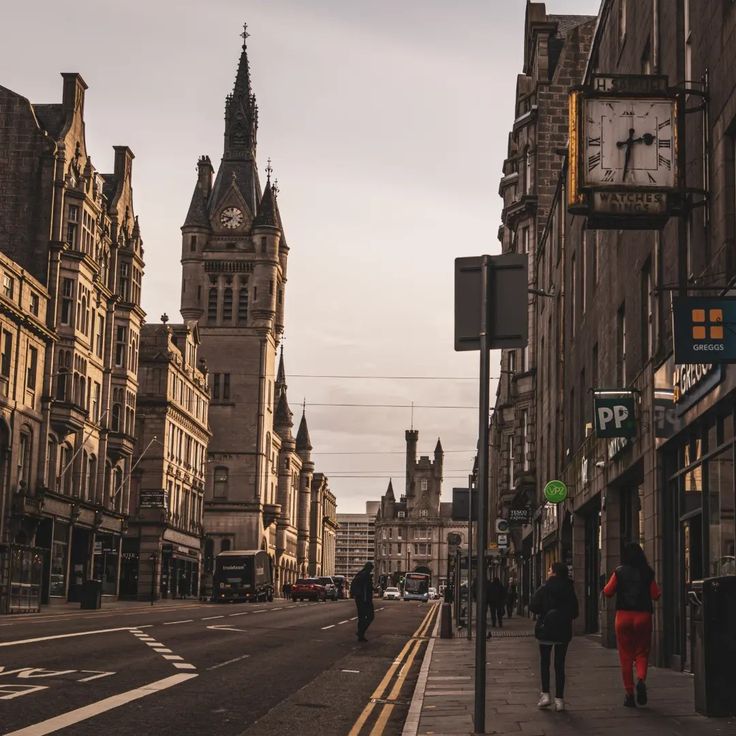 two people walking down the street in front of tall buildings with clock tower on top