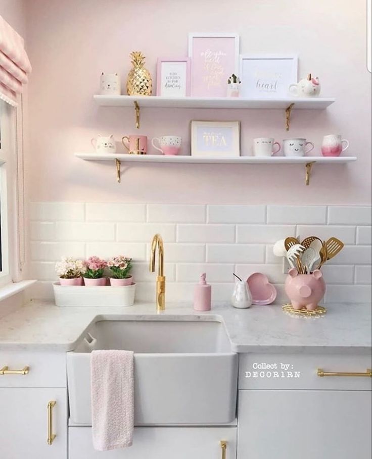 a white kitchen with pink walls and gold accessories on the shelf above the sink,