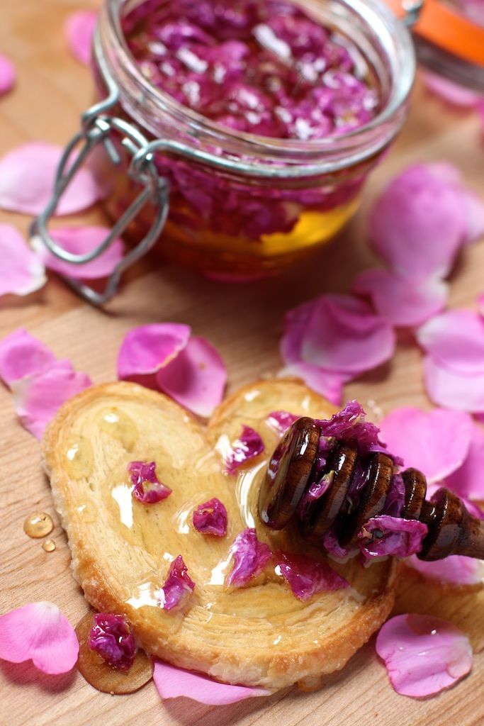 a jar of honey sits on top of a piece of bread with pink petals scattered around it