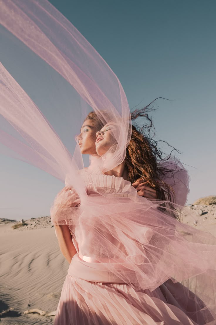 two women in pink dresses and veils standing on sand dunes with their arms around each other