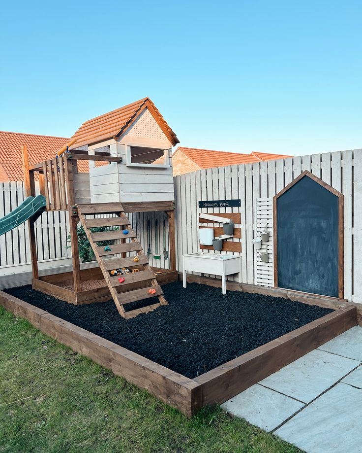 a backyard play area with a wooden swing set and black gravel in the ground next to a white picket fence