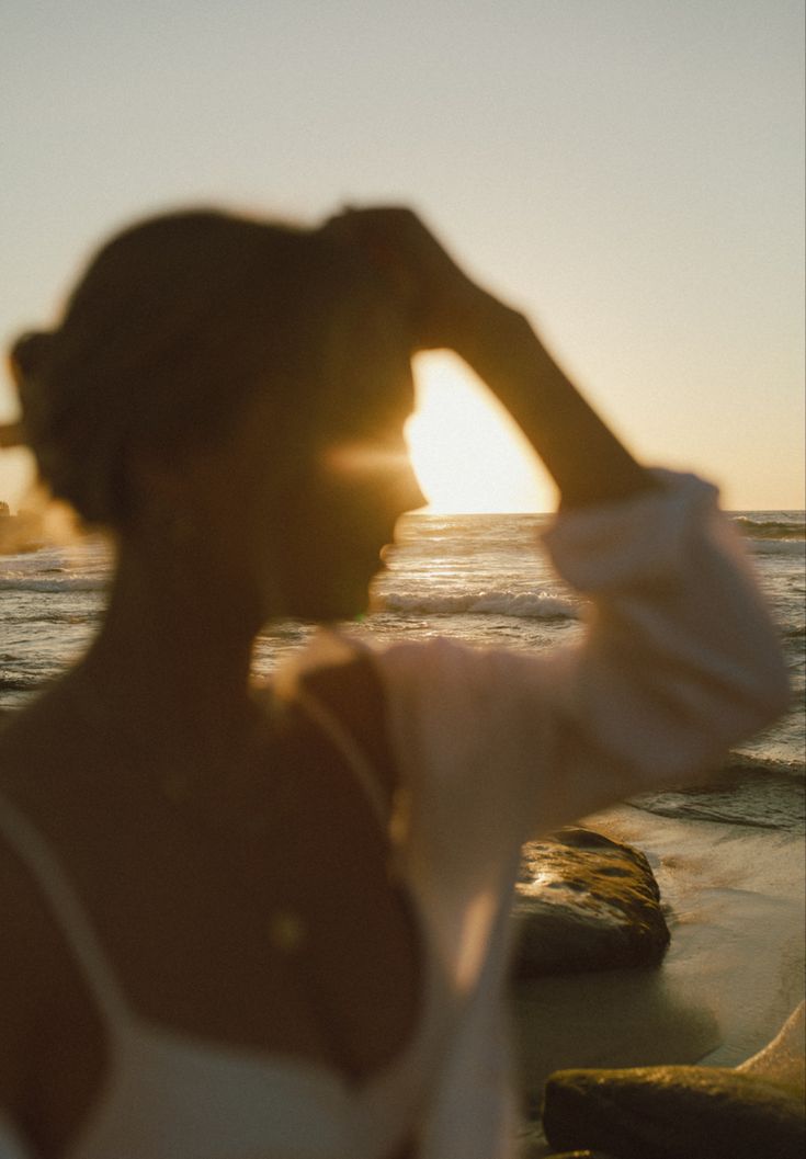 a woman standing on top of a beach next to the ocean holding her hair up