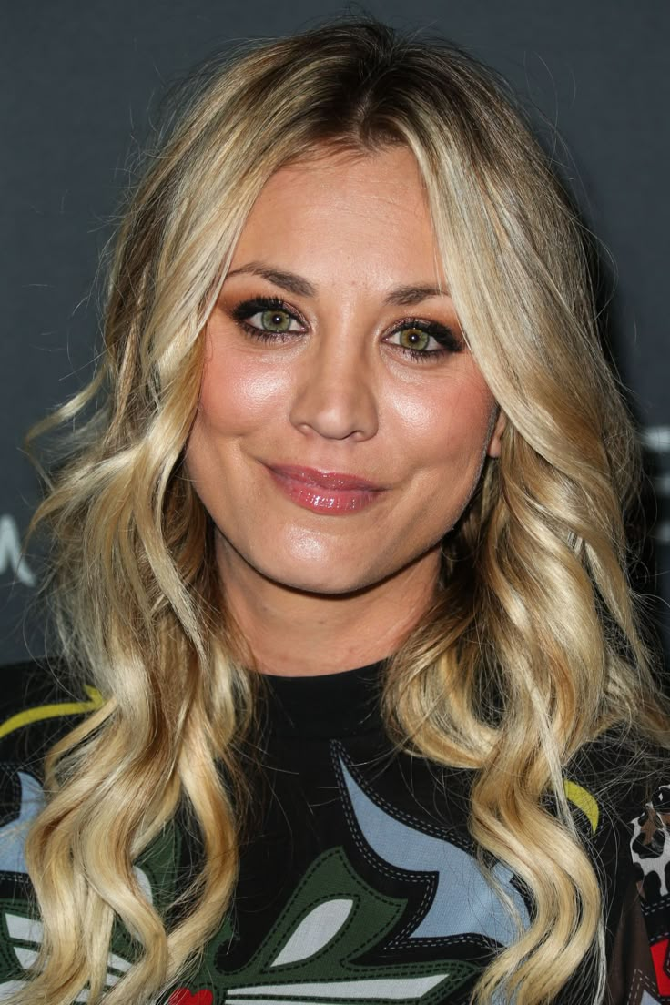 a woman with long blonde hair and green eyeshadow smiles at the camera while standing in front of a black background