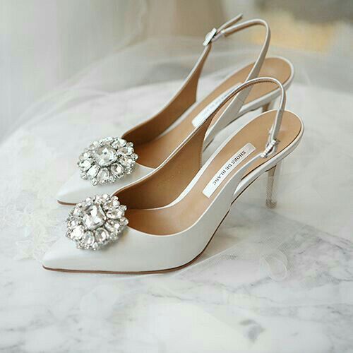 a pair of white high heel shoes with jeweled buckles on the toes, sitting on a marble surface