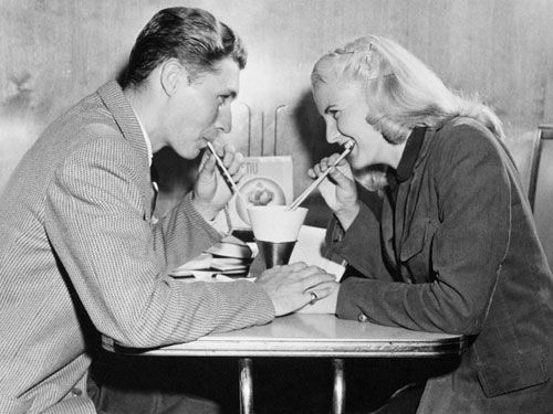 two people sitting at a table eating food and drinking from cups with chopsticks in their mouths