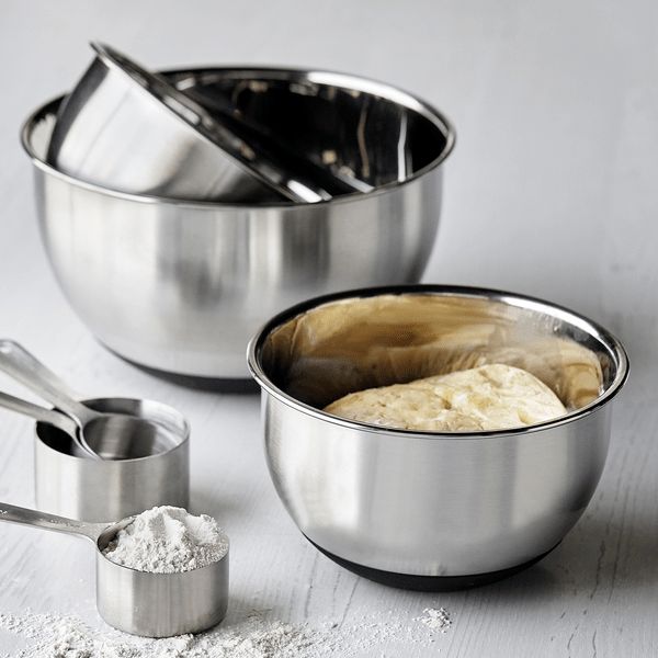 three stainless steel mixing bowls and measuring spoons on a white wooden table with flour in them