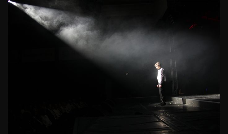 a man standing in the middle of a stage with his hands on his hips and light coming from behind him