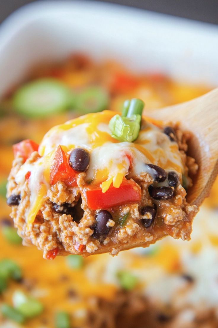 a wooden spoon full of taco meat and black beans with cheese on top, in a casserole dish